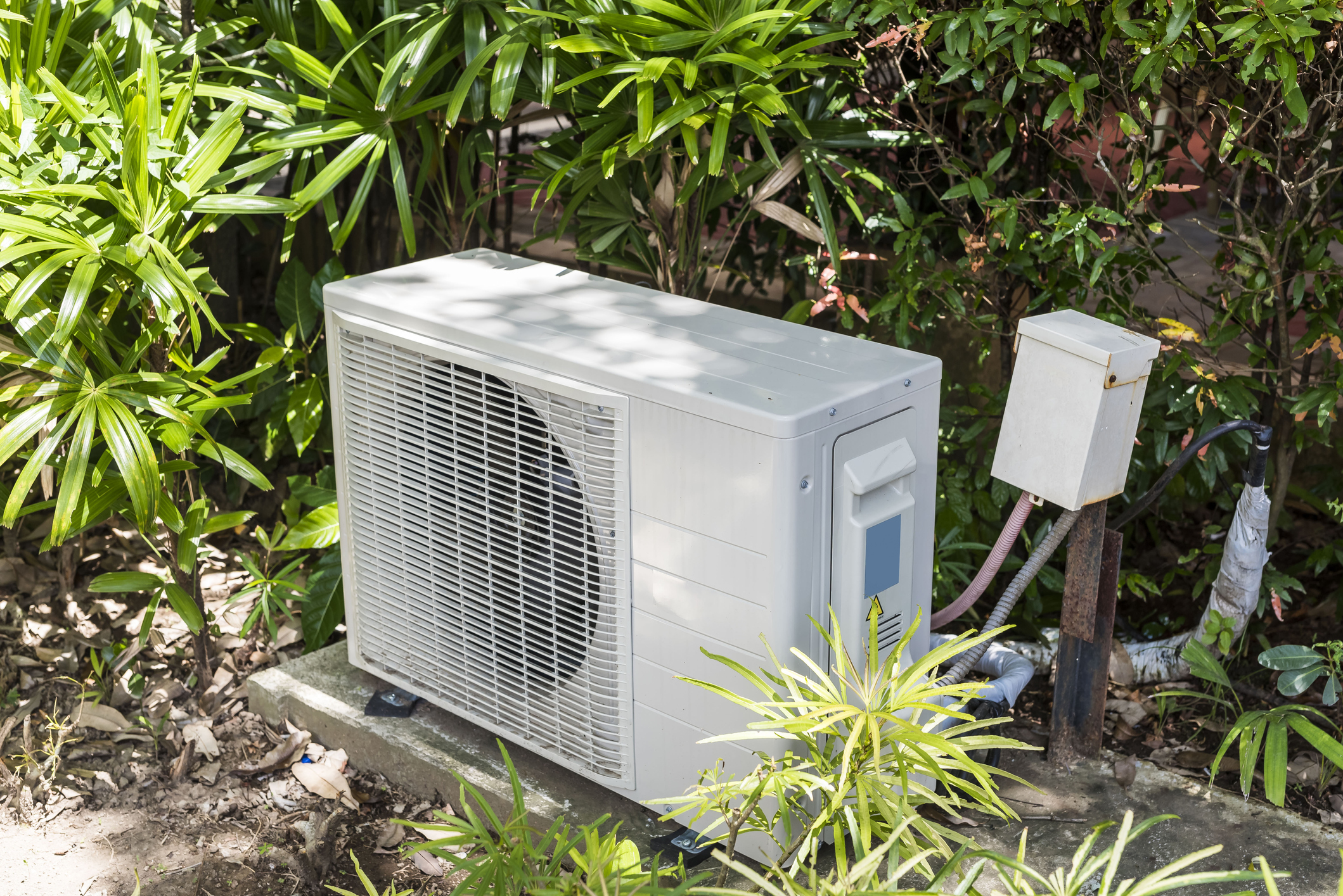 A Split Type Inverter Air Conditioning Condenser Mounted Outside A House Partially Concealed With Shrubs. Hvac For Tropical Countries.